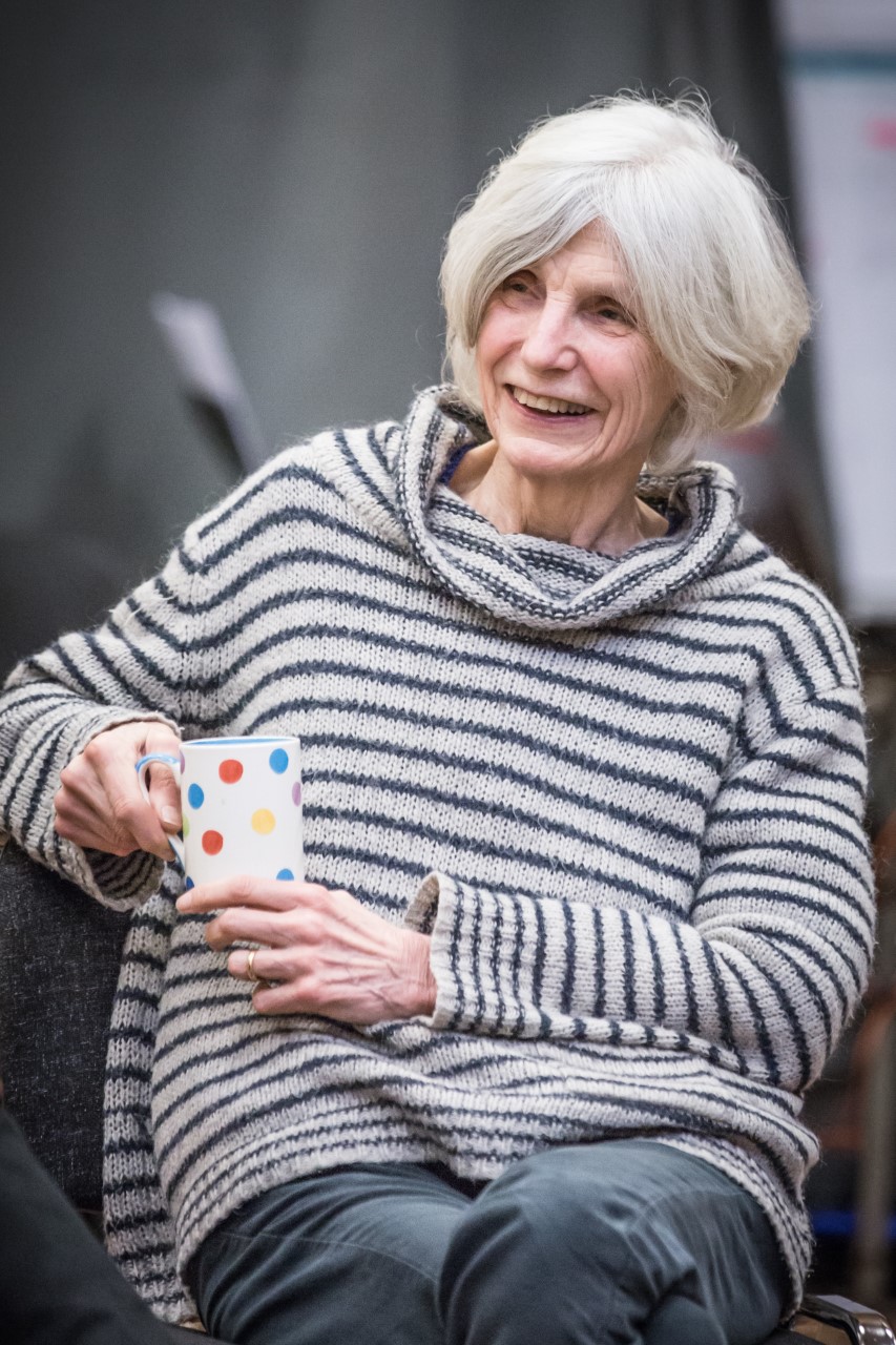 Caryl Churchill sits comfortably on a chair in a collaborative space, wearing a grey and blue striped jumper and holding a white mug with brightly coloured spots.